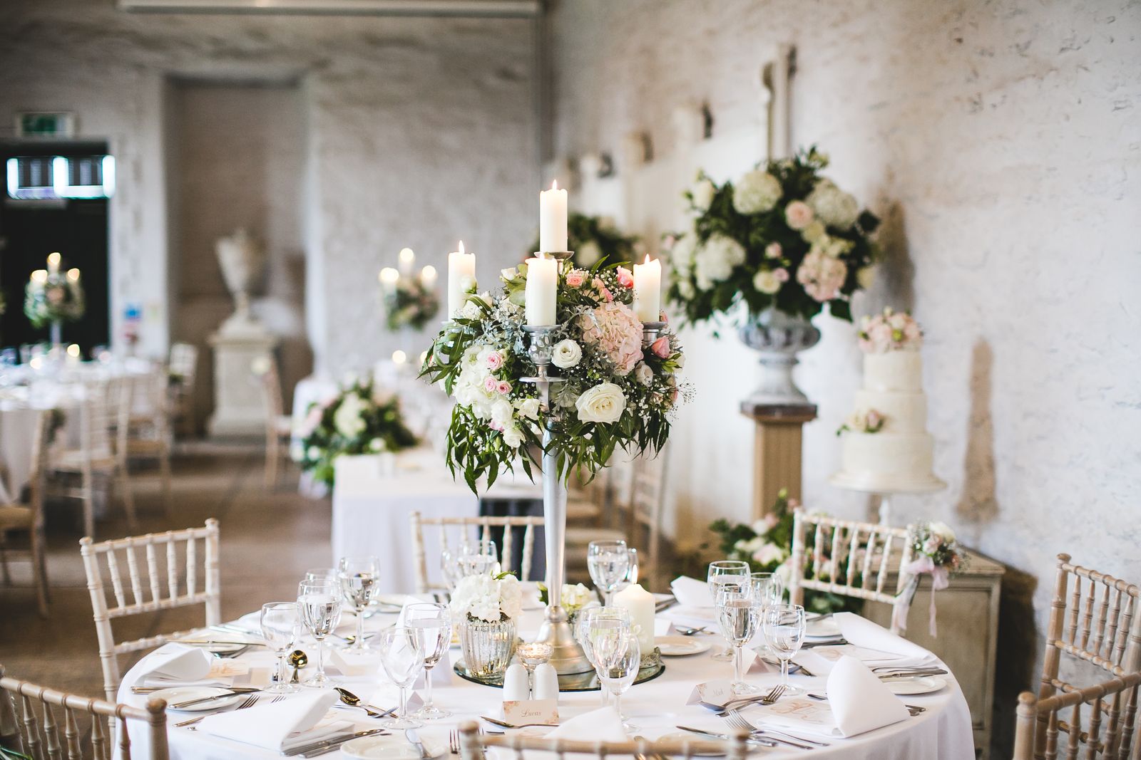 Tall floral arrangements with pillar candles in front of white rustic stone wall.