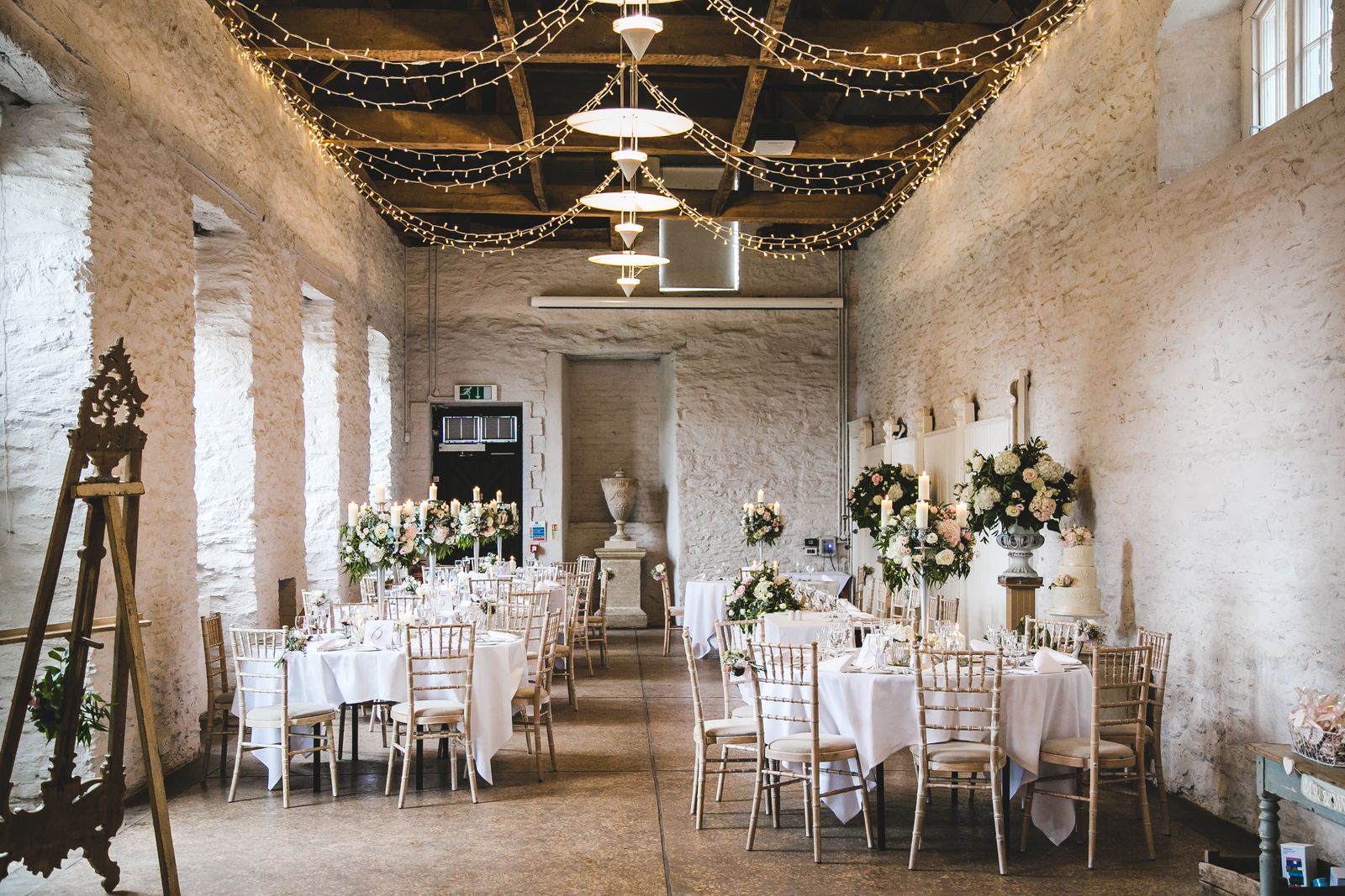 Stone barn style building with fairy light canopy, timber beams across the ceiling and romantic wedding tables.