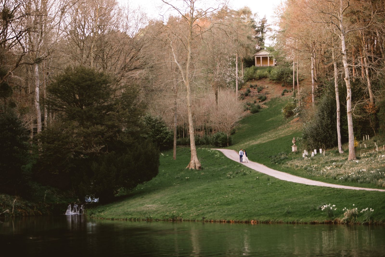 Couple walk along the lake path with the temple behind - the setting for outdoor wedding ceremonies in Somerset at Hestercombe Gardens